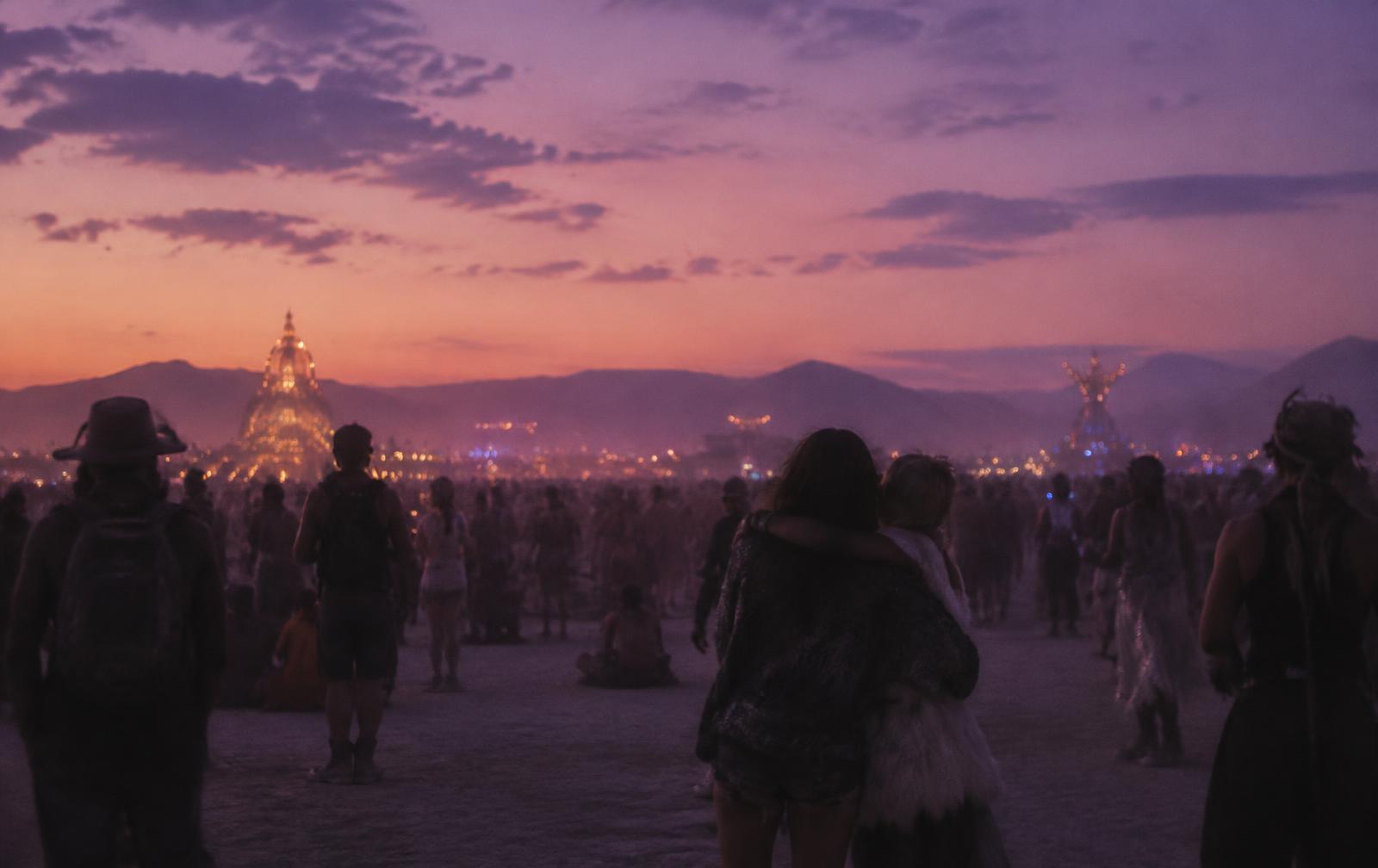 A festival crowd at dusk, two friends holding each other in the middle of it